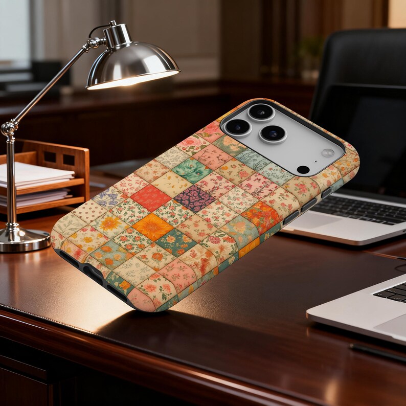 May include: A phone case with a patchwork quilt design. The case features a pattern of small, colorful squares with floral and geometric prints in shades of orange, pink, green, and cream. The case is shown on a dark wood desk.