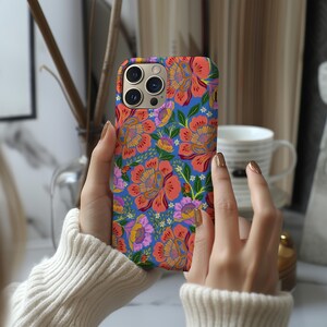 May include: A phone case with a vibrant floral pattern. The case features large orange and pink flowers with yellow centers, set against a blue background. Green leaves and smaller purple flowers complete the design. The case has a camera cutout.