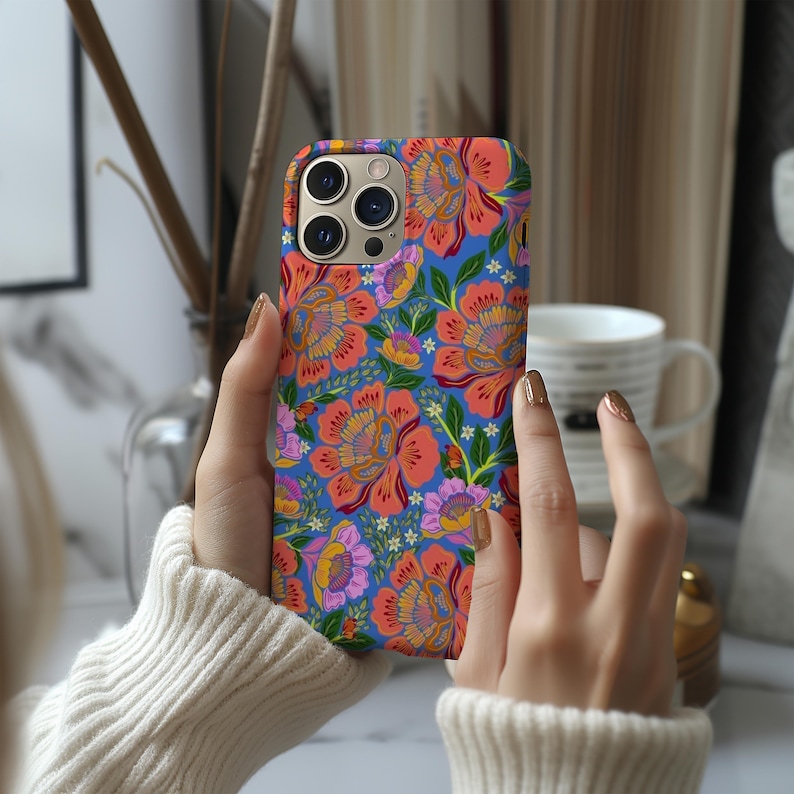 May include: A phone case with a vibrant floral pattern. The case features large orange and pink flowers with yellow centers, set against a blue background. Green leaves and smaller purple flowers complete the design. The case has a camera cutout.