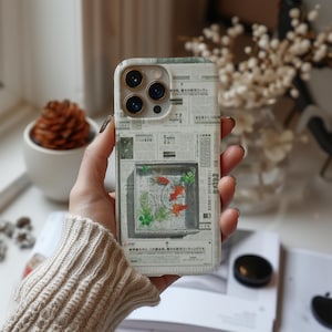 May include: A phone case with a newspaper print design. The case features a rectangular image of a fish tank with orange fish and green plants. The case has a camera cutout and is held by a person.