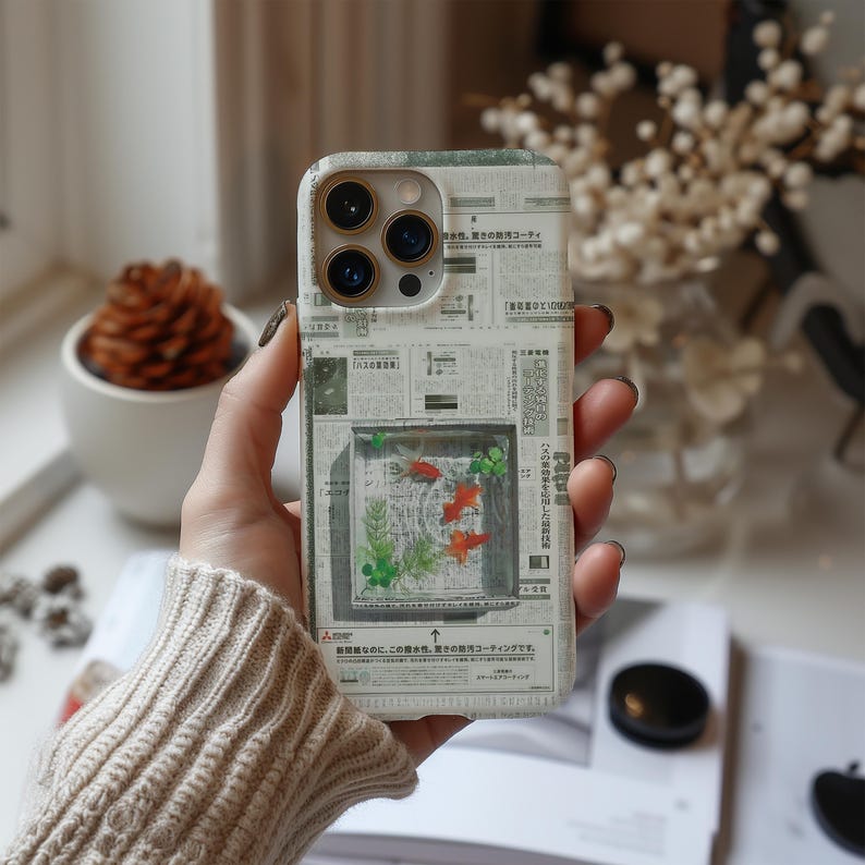 May include: A phone case with a newspaper print design. The case features a rectangular image of a fish tank with orange fish and green plants. The case has a camera cutout and is held by a person.