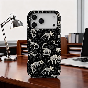 May include: Black phone case featuring a repeating pattern of white dinosaur skeletons. The design includes various dinosaur species and fossil-like elements. The case is shown on a wooden desk with a desk lamp and laptop in the background.