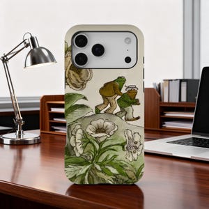 May include: A phone case featuring a whimsical illustration of two anthropomorphic frogs in clothing, walking on a hill. The design includes flowers and foliage in a vintage style, with a cream-colored background.