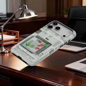 May include: A smartphone case featuring a unique design with a fish tank motif. The case has a newspaper print background and a rectangular window displaying orange fish and green plants. The case is on a dark wooden desk.