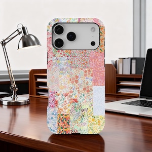 May include: A phone case with a patchwork design featuring various floral patterns. The case displays a mix of pink, red, yellow, and blue floral prints. It rests on a wooden desk, next to a silver desk lamp.