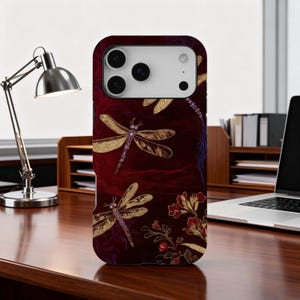 May include: A burgundy phone case featuring a pattern of gold dragonflies and floral accents. The case has a textured appearance and is designed to fit a smartphone. The design includes a mix of botanical and insect elements.