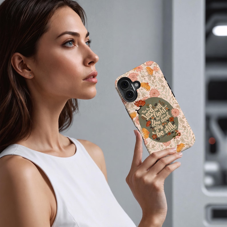 May include: A phone case with a floral pattern in peach, orange, and green. The case features the text "The Lord will fight for you, you need only be still" in a stylized font. The case is held by a person.