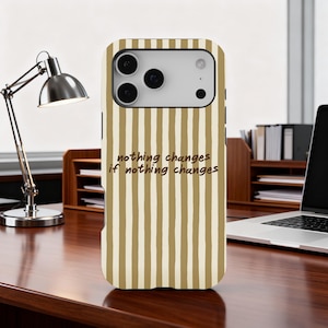Puede incluir: Funda de teléfono con un patrón de rayas verticales en beige y blanco. La funda presenta el texto "nothing changes if nothing changes" en un estilo manuscrito. La funda está sobre un escritorio de madera.