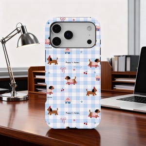 Puede incluir: Funda de teléfono con un patrón de cuadros azul y blanco con ilustraciones de perros salchicha, casas, lazos, estrellas y cerezas. El texto "Puppy's home" se repite. La funda tiene un recorte para la cámara y está sobre un escritorio de madera.