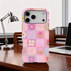 May include: A pink phone case with a floral design. The case has a grid of squares, each with a different flower in pink, purple, and yellow. The case is on a wooden desk.