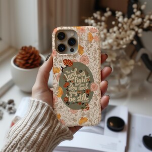 May include: A floral patterned phone case in cream, peach, and yellow. The case features a quote in a green oval: "The Lord will fight for you. You need only be still."
