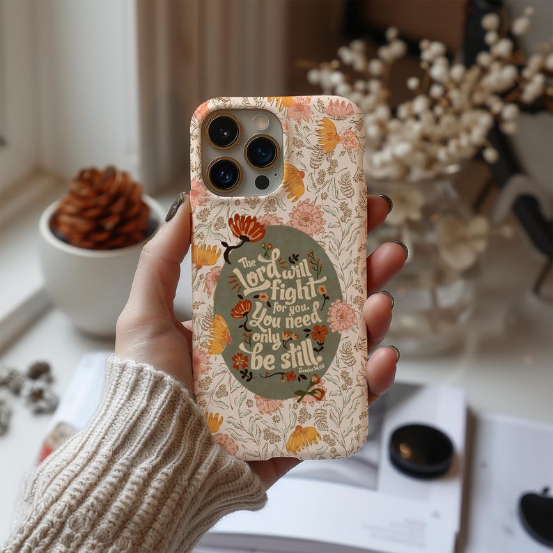 May include: A floral patterned phone case in cream, peach, and yellow. The case features a quote in a green oval: "The Lord will fight for you. You need only be still."