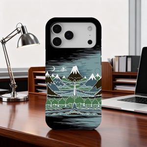 Puede incluir: Funda de teléfono negra con un diseño de paisaje con montañas, árboles y agua. La obra de arte utiliza tonos verdes, azules y blancos, con un borde negro. La funda está sobre un escritorio de madera.