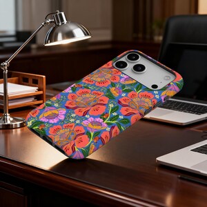 May include: A phone case with a vibrant floral pattern. The design features large, stylized flowers in shades of red, orange, and purple against a blue background. Green leaves and small white flowers complete the design. The case is shown on a dark wooden desk.