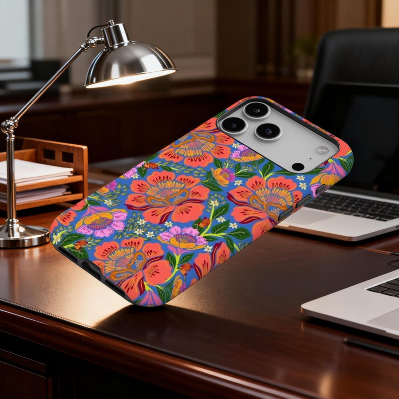 May include: A phone case with a vibrant floral pattern. The design features large, stylized flowers in shades of red, orange, and purple against a blue background. Green leaves and small white flowers complete the design. The case is shown on a dark wooden desk.