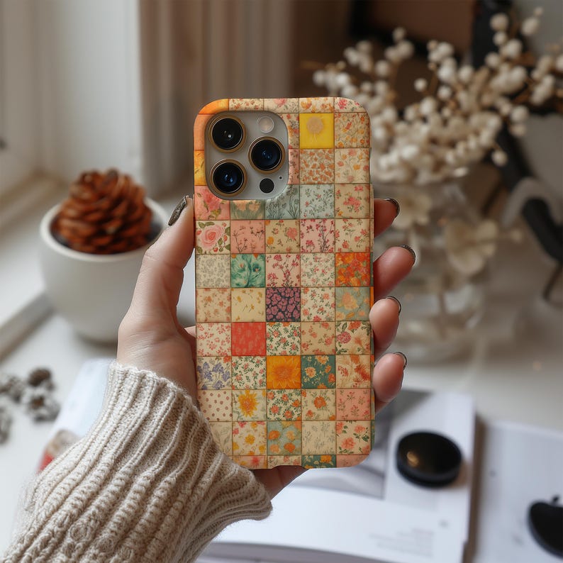 May include: A floral patchwork design phone case. The case features a variety of small, square patterns with different floral prints in shades of pink, yellow, orange, and green. The phone case is held in a person's hand.