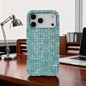 Könnte beinhalten: Eine türkisfarbene Handyhülle mit einem Mosaikfliesenmuster. Die Hülle hat ein quadratisches Rasterdesign mit glänzendem Finish. Die Handyhülle liegt auf einem Holzschreibtisch mit einer silbernen Schreibtischlampe und einem Laptop im Hintergrund.