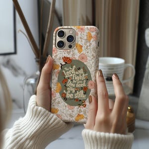 May include: A floral phone case with a cream background and orange and pink flowers. The case features a green oval with the text "The Lord will fight for you. You need only be still."