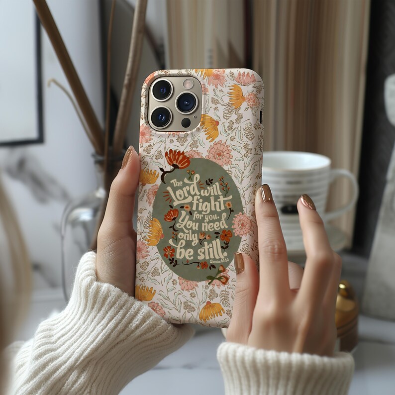May include: A floral phone case with a cream background and orange and pink flowers. The case features a green oval with the text "The Lord will fight for you. You need only be still."