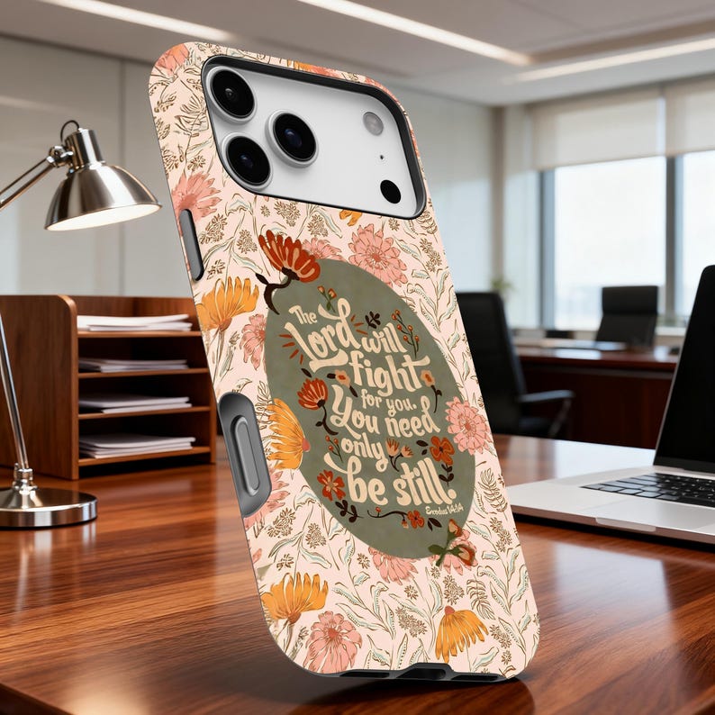 May include: A phone case with a floral pattern in peach, orange, and pink hues. The case features the text "The Lord will fight for you. You need only be still." in a decorative oval. The case is on a wooden desk.