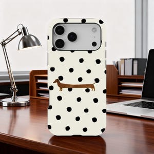 Puede incluir: Una funda de teléfono con fondo crema y lunares negros. Una ilustración de perro salchicha marrón está centrada en la funda. La funda del teléfono está sobre un escritorio de madera con una lámpara de escritorio plateada y un portátil.