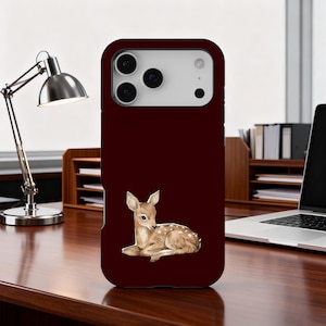 May include: A maroon phone case with a watercolour illustration of a fawn with white spots. The fawn is lying down, with its head turned slightly to the side. The case has a glossy finish and a camera cutout.