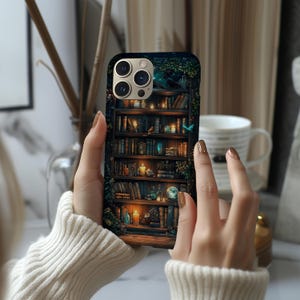 May include: A phone case featuring a detailed illustration of a bookshelf filled with books, candles, and decorative items. The case has a dark background with a touch of green foliage. The phone case is held by a person wearing a white sweater.
