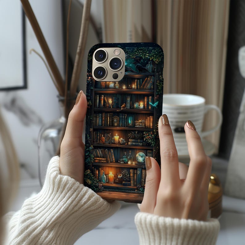 May include: A phone case featuring a detailed illustration of a bookshelf filled with books, candles, and decorative items. The case has a dark background with a touch of green foliage. The phone case is held by a person wearing a white sweater.