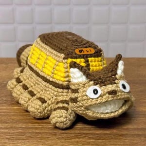Puede incluir: Juguete de crochet con forma de personaje de dibujos animados. Tiene un cuerpo beige con rayas marrones, una espalda amarilla y marrón, y una cara sonriente con ojos grandes. El texto japonés "めい" está en una pequeña etiqueta.