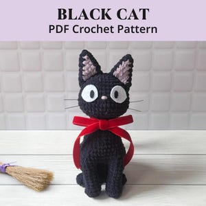 Puede incluir: Un muñeco de gato tejido a ganchillo negro con grandes ojos de fieltro blanco, una pequeña nariz rosa y largos bigotes. El gato tiene orejas puntiagudas con detalles en rosa claro y lleva una cinta de terciopelo rojo atada al cuello. Texto: BLACK CAT PDF Crochet Pattern.