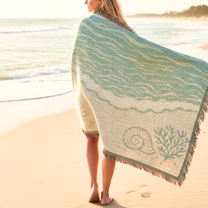 Peut inclure: Une couverture de plage aux tons de bleu sarcelle et crème, ornée d'un motif de vagues. Elle présente un coquillage et du corail, avec des bords frangés. La personne porte la couverture sur une plage de sable.