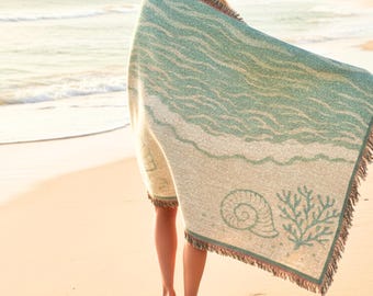 May include: A beach-themed blanket with a wave pattern in shades of teal and cream. The blanket features a seashell and coral design, with fringed edges. The person in the image is wearing the blanket on a sandy beach.