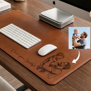 Puede incluir: Un tapete de escritorio de cuero marrón con un teclado y un ratón blancos. El tapete tiene el texto "I love you, Daddy!" y un boceto de un padre y un niño. Un monitor de computadora y una foto también están en el escritorio.