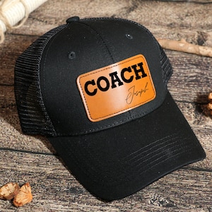 May include: Black trucker hat with a mesh back and a curved brim. The hat features a brown leather patch with the word "COACH" in bold black letters and the name "Joseph" in a script font.