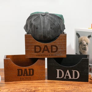 以下が含まれることがあります： 前面に「DAD」の文字が刻まれた3つの木箱。1つの箱には野球帽が入っています。箱は茶色と黒のさまざまな色合いです。1つの箱には「Andy & Steve & Nicole」というテキストがあり、もう1つには「Est. 2025」というテキストがあります。