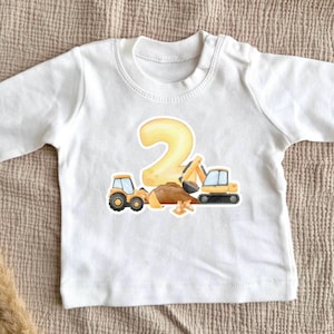 Op de afbeelding: Witte baby-shirt met lange mouwen met een kleurrijke illustratie. Het ontwerp toont een groot geel nummer 2, twee bouwmachines en een hoop aarde. Het shirt is gemaakt van zacht materiaal.