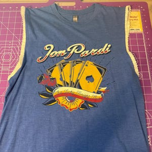 Puede incluir: Camiseta sin mangas azul con el texto "Jon Pardi" en letras doradas, con un gráfico de naipes y una pancarta que dice "Be Lucky Tonight Tour". La camiseta sin mangas tiene un ribete de encaje.