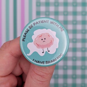 May include: A round teal pin-back button featuring a central illustration of a sad pink brain character surrounded by a white cloud shape. The text printed in dark grey around the edge reads, PLEASE BE PATIENT WITH ME I HAVE BRAIN FOG.