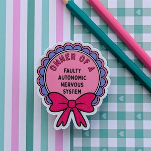 May include: A circular sticker shaped like a rosette with a pink center, blue scalloped border, and a bright pink bow at the bottom. The text reads OWNER OF A FAULTY AUTONOMIC NERVOUS SYSTEM in black capital letters. Two colored pencils lie nearby.