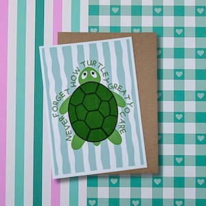 May include: A rectangular greeting card featuring a green turtle illustration centred on a light blue and white wavy striped background. The text "NEVER FORGET HOW TURTLEY GREAT YOU ARE" curves around the turtle. The card rests on a brown envelope.