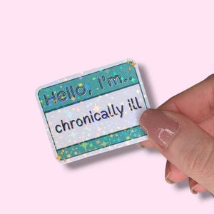 May include: A rectangular sticker with the text "Hello, I'm.. chronically ill" in white and teal. The sticker has a holographic, glittery effect with star accents and is held by a hand against a pink background.