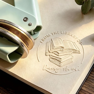 Pode incluir: Uma ferramenta de relevo em metal verde menta claro repousa sobre uma página, criando um design circular em relevo com uma pilha de livros abertos e o texto "FROM THE LIBRARY OF Daisy Arole" pressionado no papel com detalhes claros e elevados.