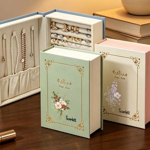 May include: Three book-shaped jewelry storage boxes in light blue, sage green, and cream. The open blue box features hooks for necklaces and padded slots for rings. The green and cream boxes are closed, displaying floral graphics and the text Scarlett.