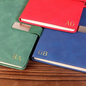 Op de afbeelding: Een verzameling gepersonaliseerde notitieboeken in groen, rood en blauw. Elk notitieboek heeft gouden initialen en een zilveren metalen sluiting. De notitieboeken liggen op een houten oppervlak.