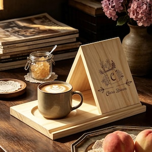 Peut inclure: Un support en bois clair en forme de A avec un motif floral gravé au laser, la lettre C et le nom Clara Brown. Le support comprend une base avec une découpe circulaire accueillant une tasse en céramique mouchetée remplie de café latte.