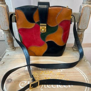 May include: A colourful patchwork leather crossbody bag with a black strap and gold-tone hardware. The bag features red, orange, green, and black leather pieces. The bag is sitting on a wooden chair.