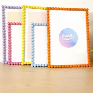 May include: Four colorful picture frames with beaded edges. The frames are in shades of lavender, pink, yellow, blue, and orange. The frames have white interiors and are displayed on a light wood surface.
