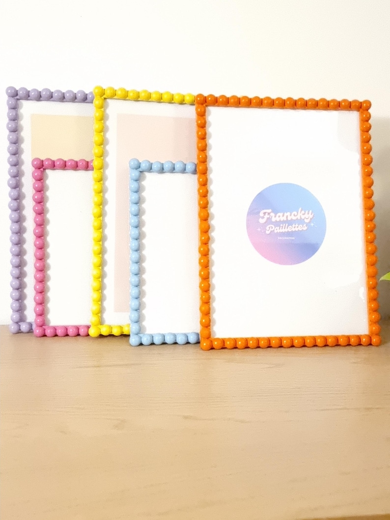 May include: Four colorful picture frames with beaded edges. The frames are in shades of lavender, pink, yellow, blue, and orange. The frames have white interiors and are displayed on a light wood surface.