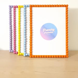 May include: A stack of colorful picture frames with rounded edges. The frames are in shades of purple, pink, yellow, blue, and orange. Each frame has a white interior. The orange frame has a sticker with the text "Francky Paillettes".