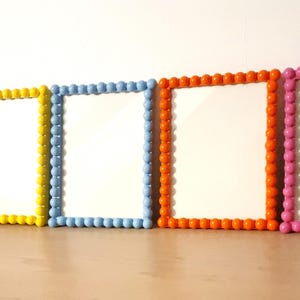 May include: Five colorful picture frames with white interiors. The frames are arranged in a row and feature a beaded border in purple, yellow, light blue, orange, and pink. The frames are on a wooden surface.